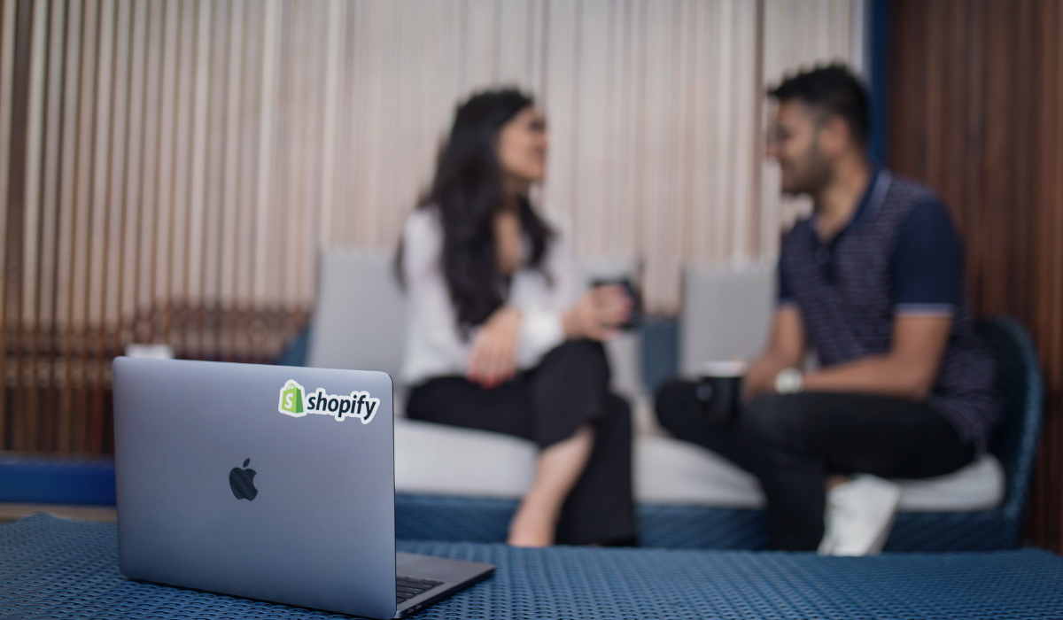 Macbook with Shopify logo, 2 people chatting in background